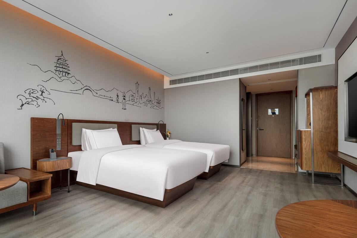 Ein Hotelzimmer mit zwei Betten, einem Holzkopfteil, einem Nachttisch, einem Fernseher und Wandgemälde.