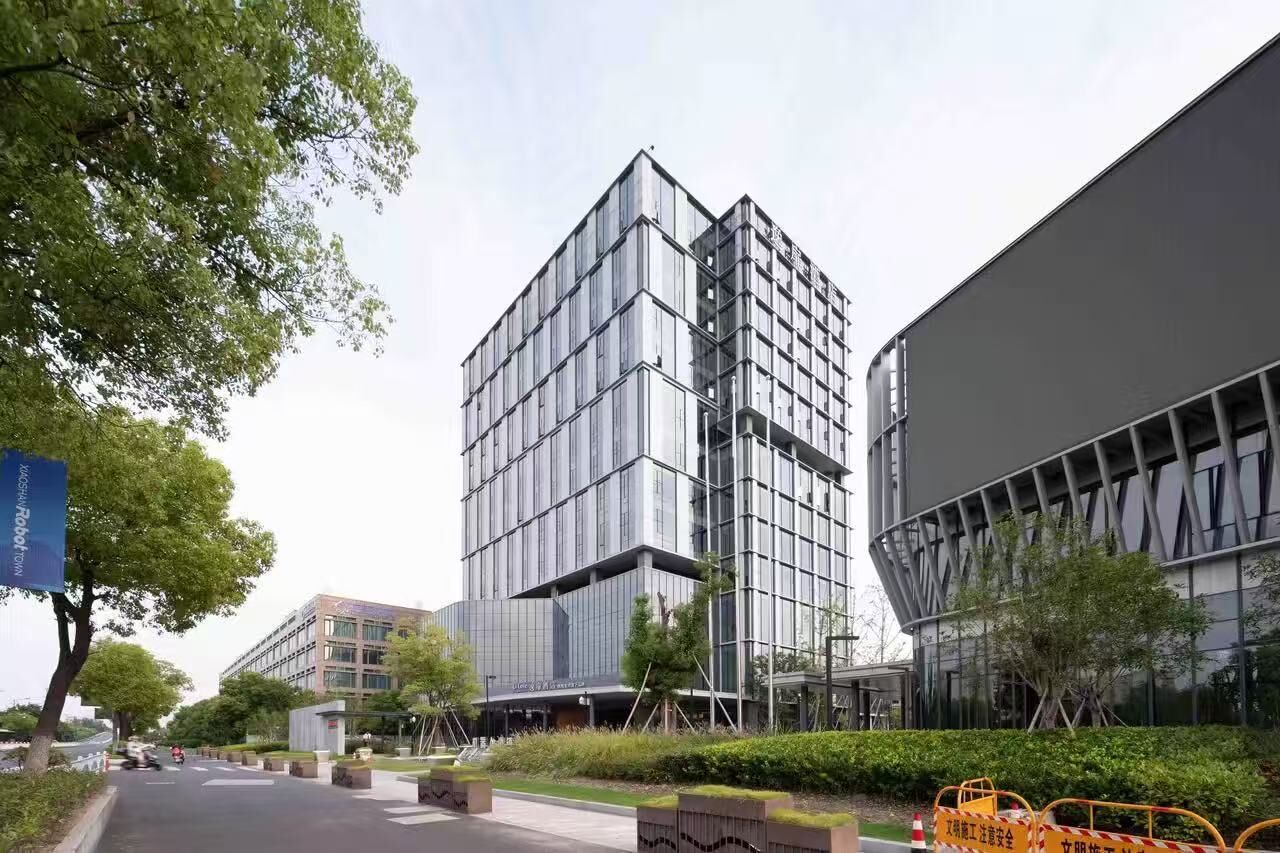 A modern building with a glass facade, surrounded by greenery and a clear sky.