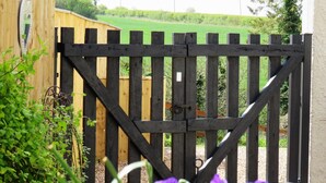 Une porte en bois avec un loquet en métal, entourée de verdure et surmontée d’une structure en bois en arrière-plan.