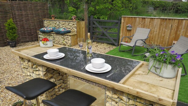Un espace bar extérieur avec un mur en pierre, deux tabourets de bar noirs, une table en bois et une clôture métallique en arrière-plan.