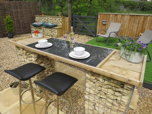An outdoor bar area with a stone wall, two black bar stools, a wooden table, and a metal fence in the background. - Private Garden Room - Rural Village (Stamford)