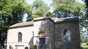 Un petit bâtiment en pierre, doté d’une porte bleue et de fenêtres en arc de cercle, entouré d’arbres et d’une zone herbeuse.
