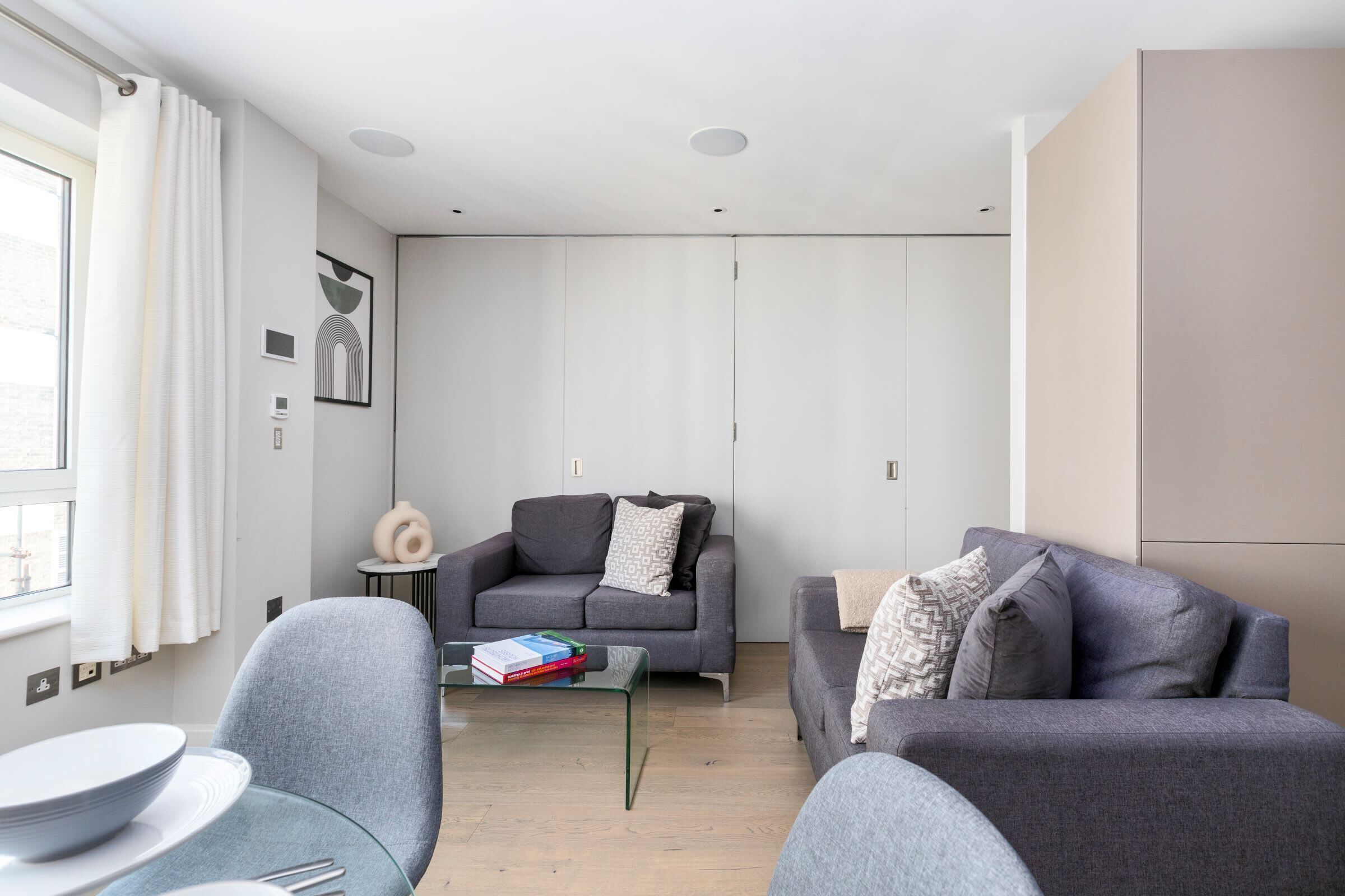 A modern living room with a grey sofa, a glass coffee table with books, a small side table, and a window with curtains.
