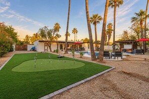A backyard with a pool, palm trees, a lawn with golf holes, and outdoor seating.