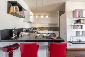 A modern kitchen with a central island, red bar stools, and built-in shelving with books and decorative items. - BnButler - Celio, 11 (Milano)
