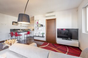 Ein modernes Wohnzimmer mit einer Couch, einem Fernseher, einer Essecke mit roten Stühlen und einer Küche im Hintergrund.