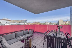 A rooftop terrace with a red wall, a grey sofa, a wooden table, and a city view. - BnButler - Celio, 11 (Milano)