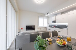 A modern kitchen with white cabinets, a dining area with a table set for two, and a TV in the background. - BnButler - De Amicis, 25 (Milano)