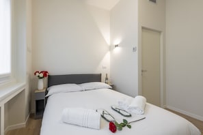 Un lit bien fait, avec du linge de lit blanc, un vase rempli de fleurs, une bouteille et une rose rouge posées sur le lit.