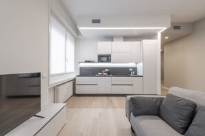 A modern kitchen with white cabinets, a microwave, and a sink. There is a grey sofa and a flat-screen TV in the living area.