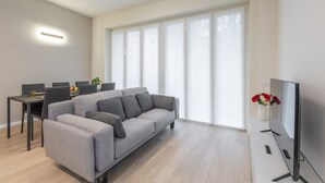 A modern living room with a grey sofa, a dining table with chairs, a flat-screen TV, and large windows. - BnButler - De Amicis, 25 (Milano)