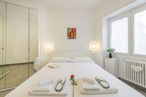 A hotel room with a bed, two bedside lamps, a radiator, a window with curtains, a small plant, and a painting on the wall. - BnButler - Washington, 83 (Milano)