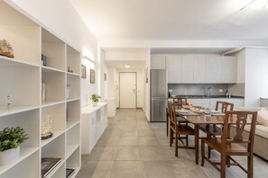 A modern kitchen with white cabinetry, a dining table with chairs, and a bookshelf with various items.