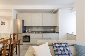 Una cocina compacta con electrodomésticos modernos, un comedor con mesa y sillas de madera, y un sofá con cojines estampados.