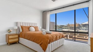 A modern hotel room with a large bed, a nightstand, a lamp, and a view of the city through large windows. - Casa Cumpari in Wannanup (Wannanup)