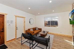 A room with a wooden door, a round mirror, a brown sofa, a black chair, a small table, and a window with a view of a building. - Willowbear Lodge By Destination Big Bear:Located in Lower Moonridge!Foosball! Close to Bear Mountain! (Big Bear Lake)