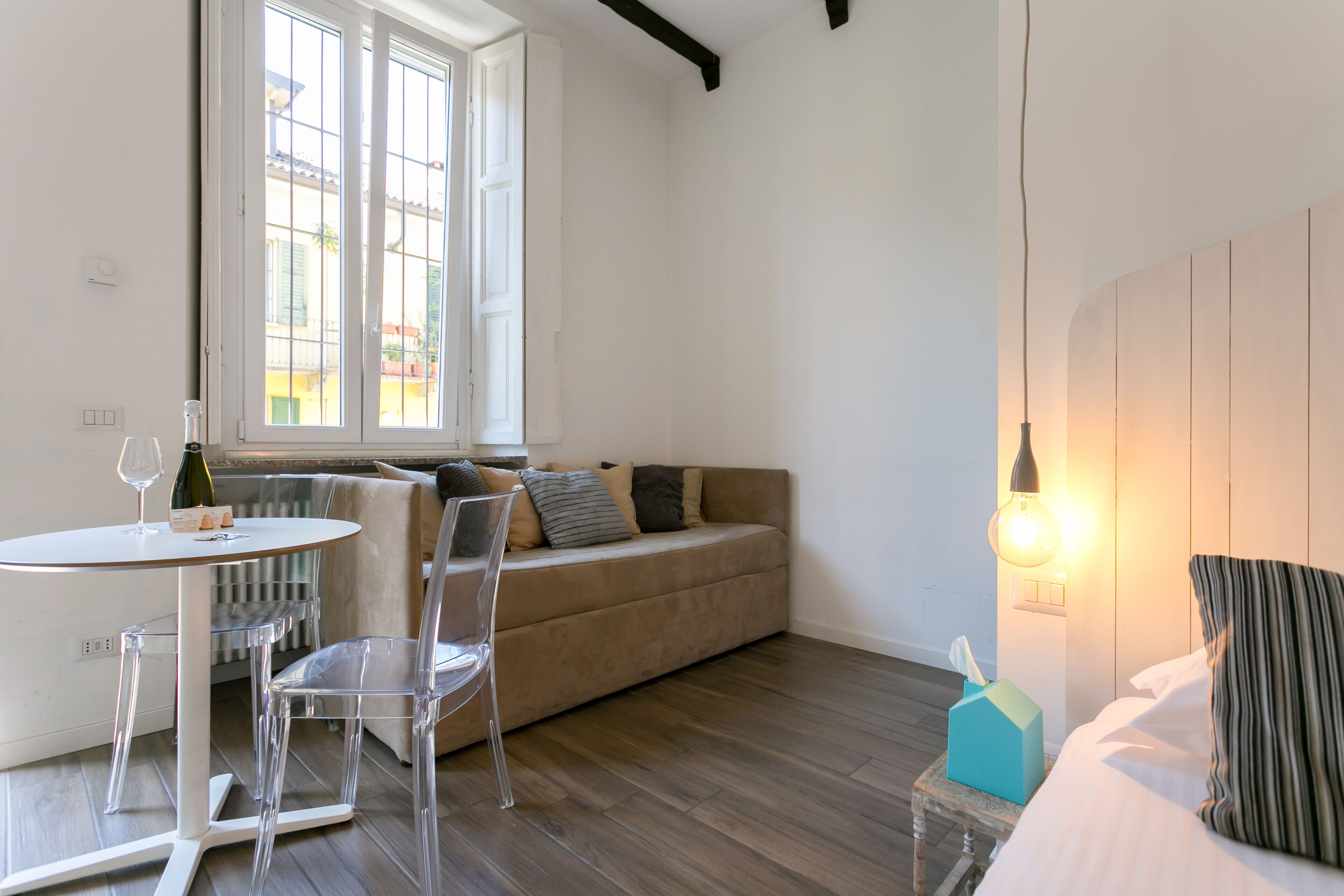 A room with a sofa, a small table with a wine glass, a chair, a bedside table with a lamp, and a radiator.