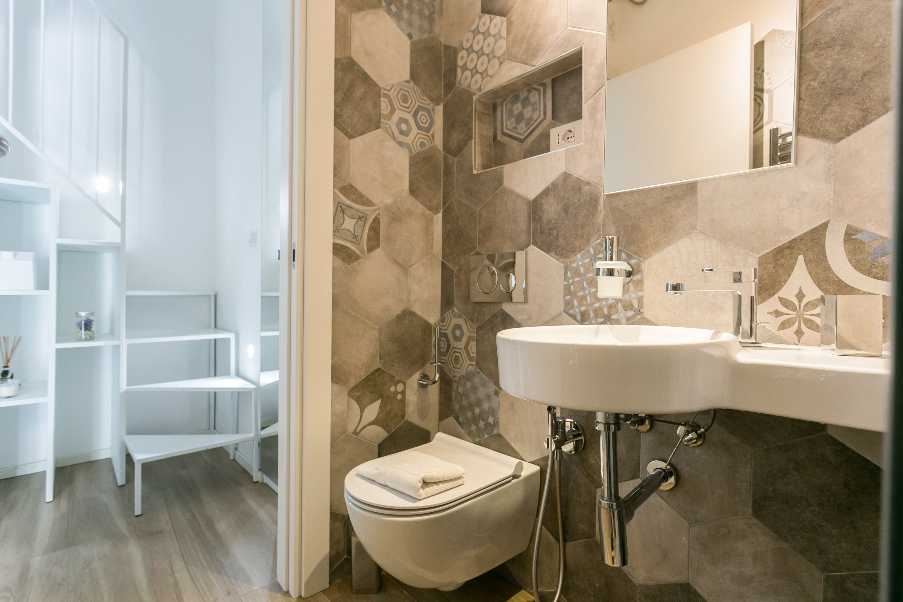 A bathroom with a toilet, sink, and shower. There are shelves and a mirror on the wall.