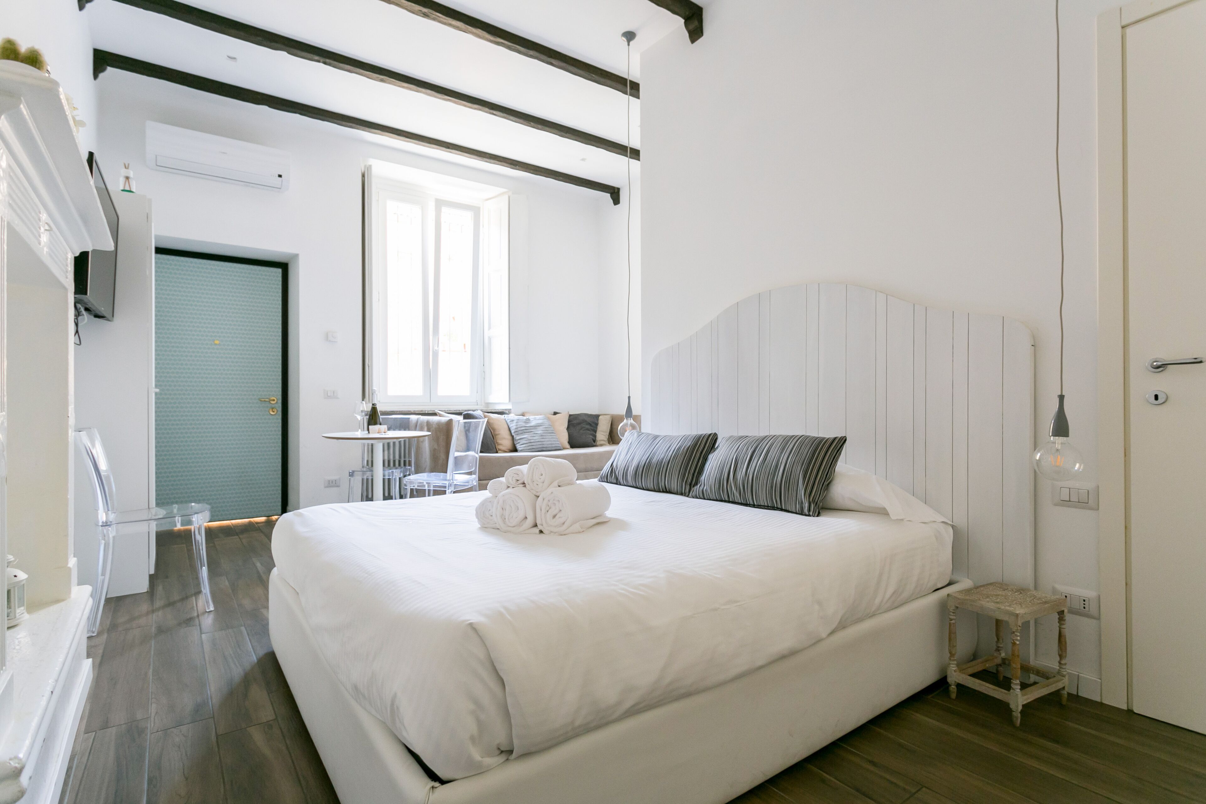 Un letto grande con lenzuola bianche e asciugamani arrotolati, un comodino di legno e una finestra con luce naturale, più una porta con maniglia.