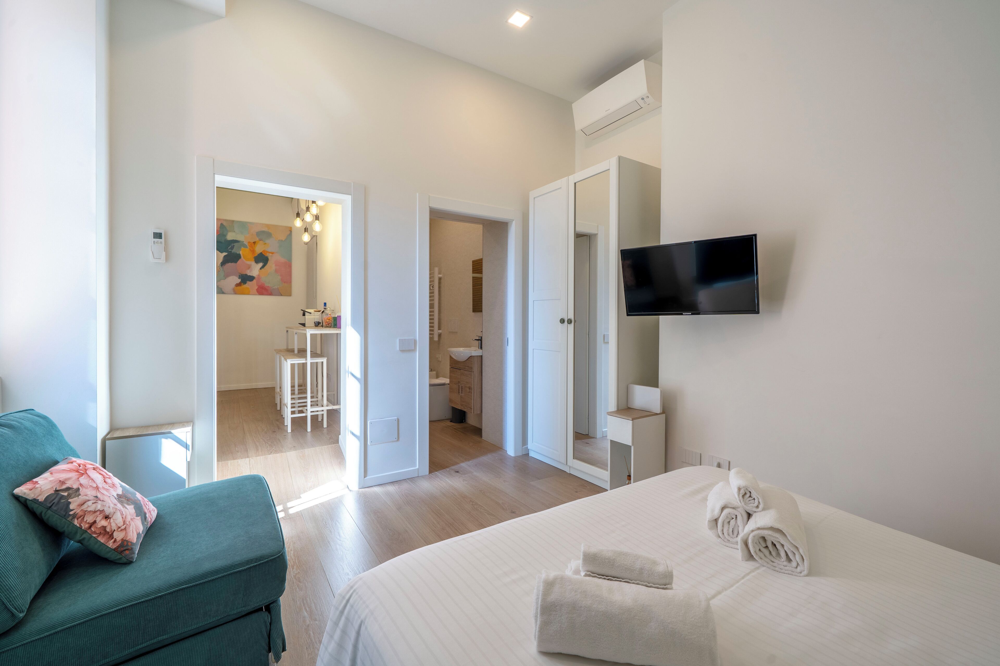 A hotel room with a bed, a TV mounted on the wall, a sofa, and a bathroom visible through an open door.
