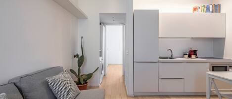 Una cocina moderna con armarios blancos, horno empotrado y microondas. Hay un sofá gris con cojines estampados y una planta en maceta.