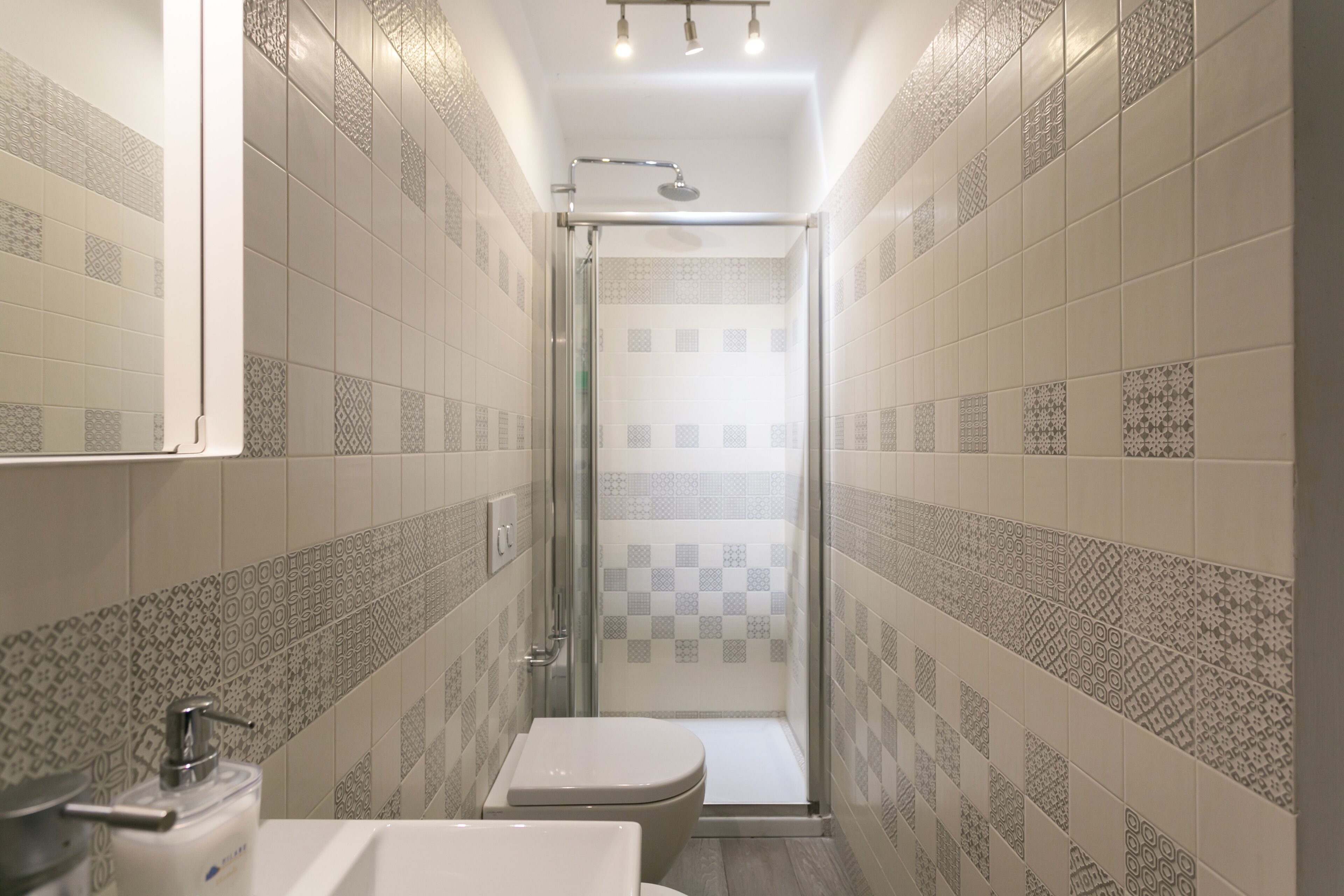 A narrow bathroom with patterned tiles, a toilet, and a glass-enclosed shower.