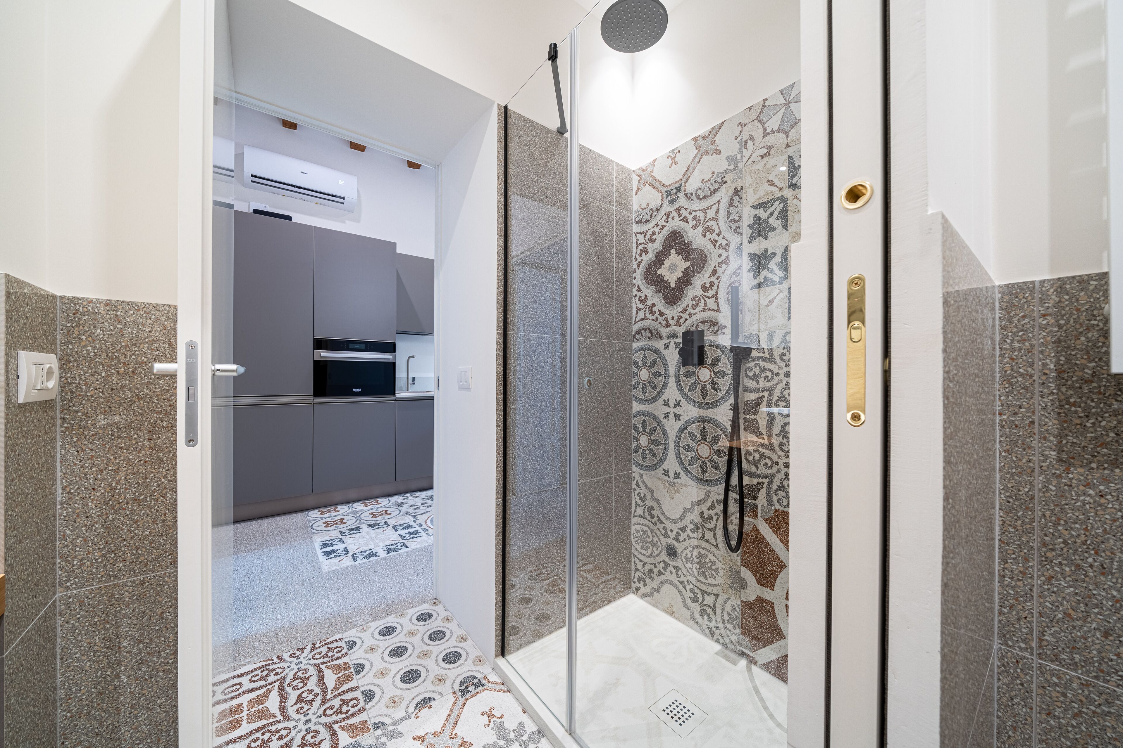 A modern bathroom with a glass shower enclosure, patterned tiles, and a visible kitchen area in the background.
