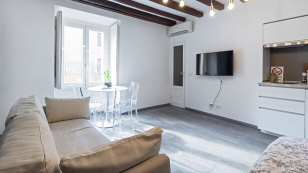 Een moderne woonkamer met een bank, een eettafel met stoelen, een aan de muur bevestigde televisie en een keuken met een aanrecht en kastjes.