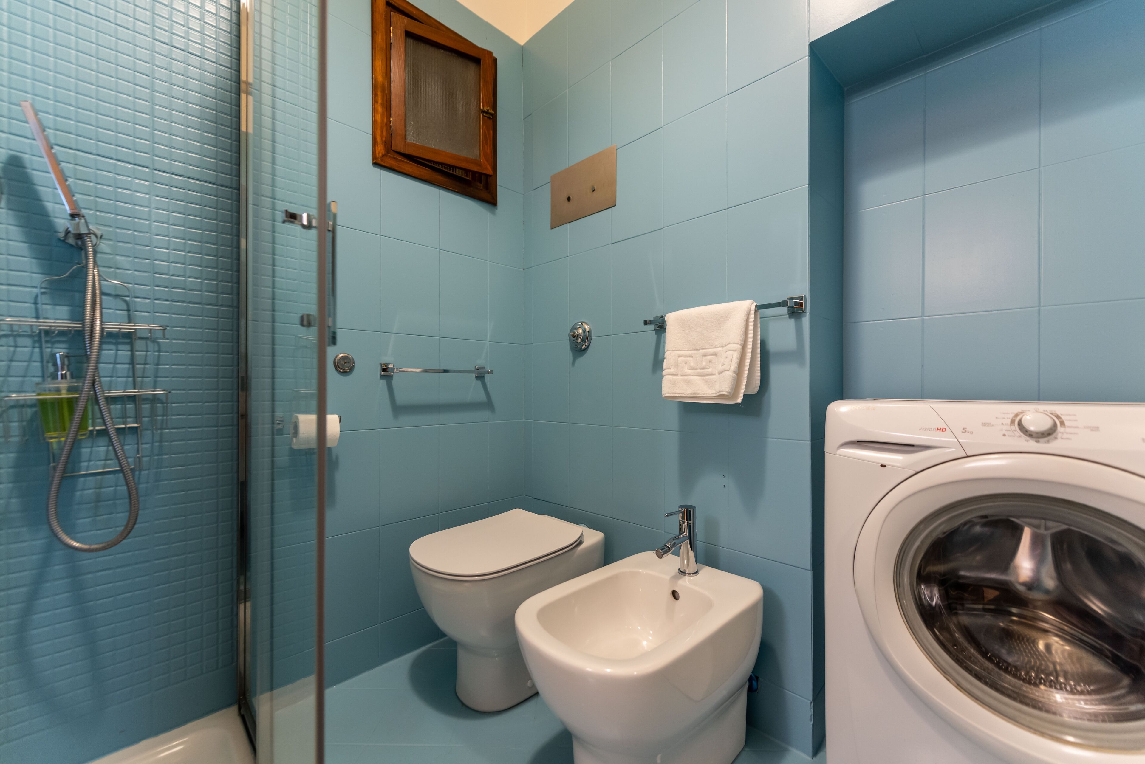 Casa de banho com cabine de duche em vidro, sanita, bidé e máquina de lavar roupa frontal.