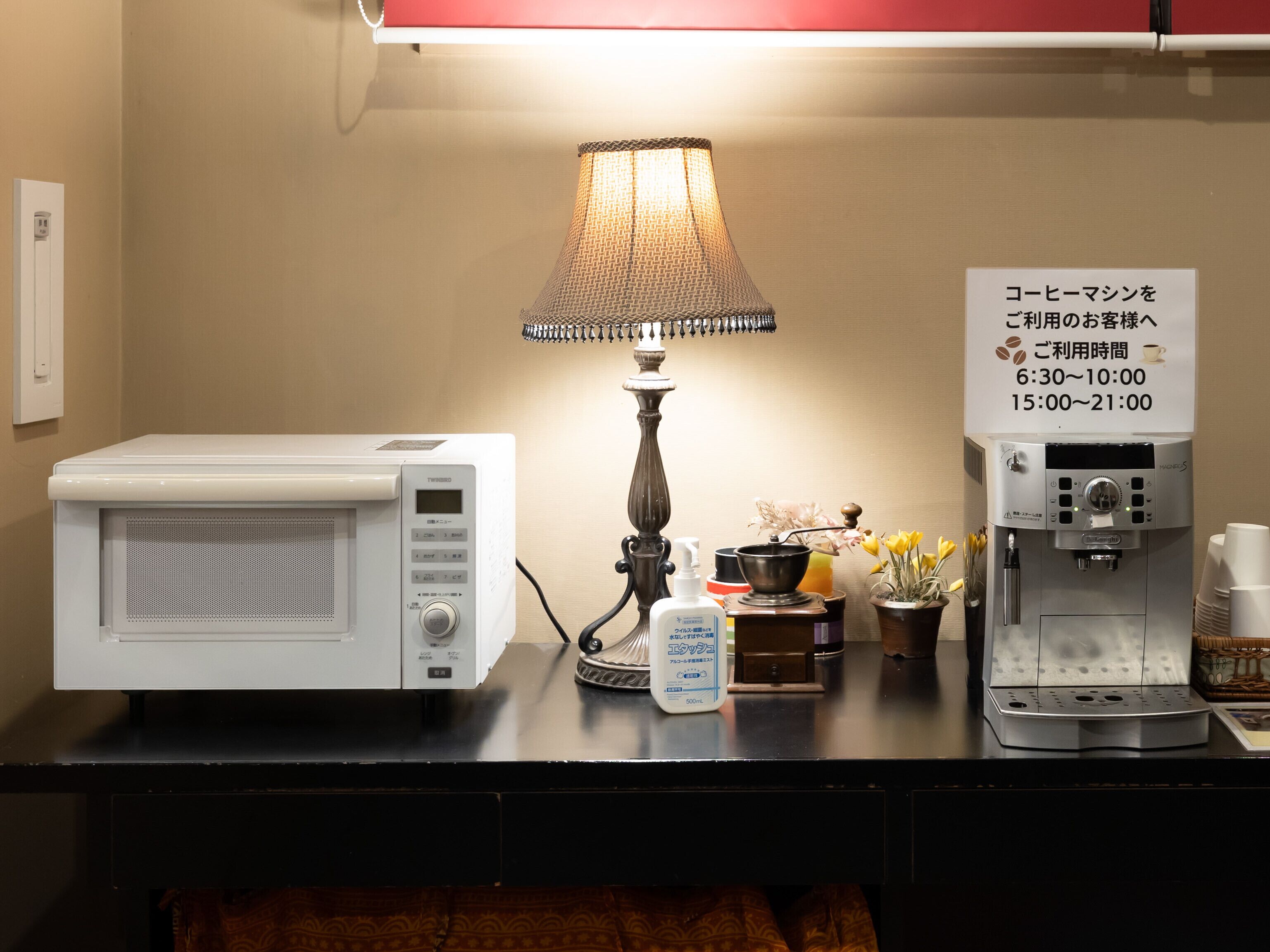 A kitchenette with a microwave, a coffee machine, a lamp, and a sign indicating coffee shop hours.