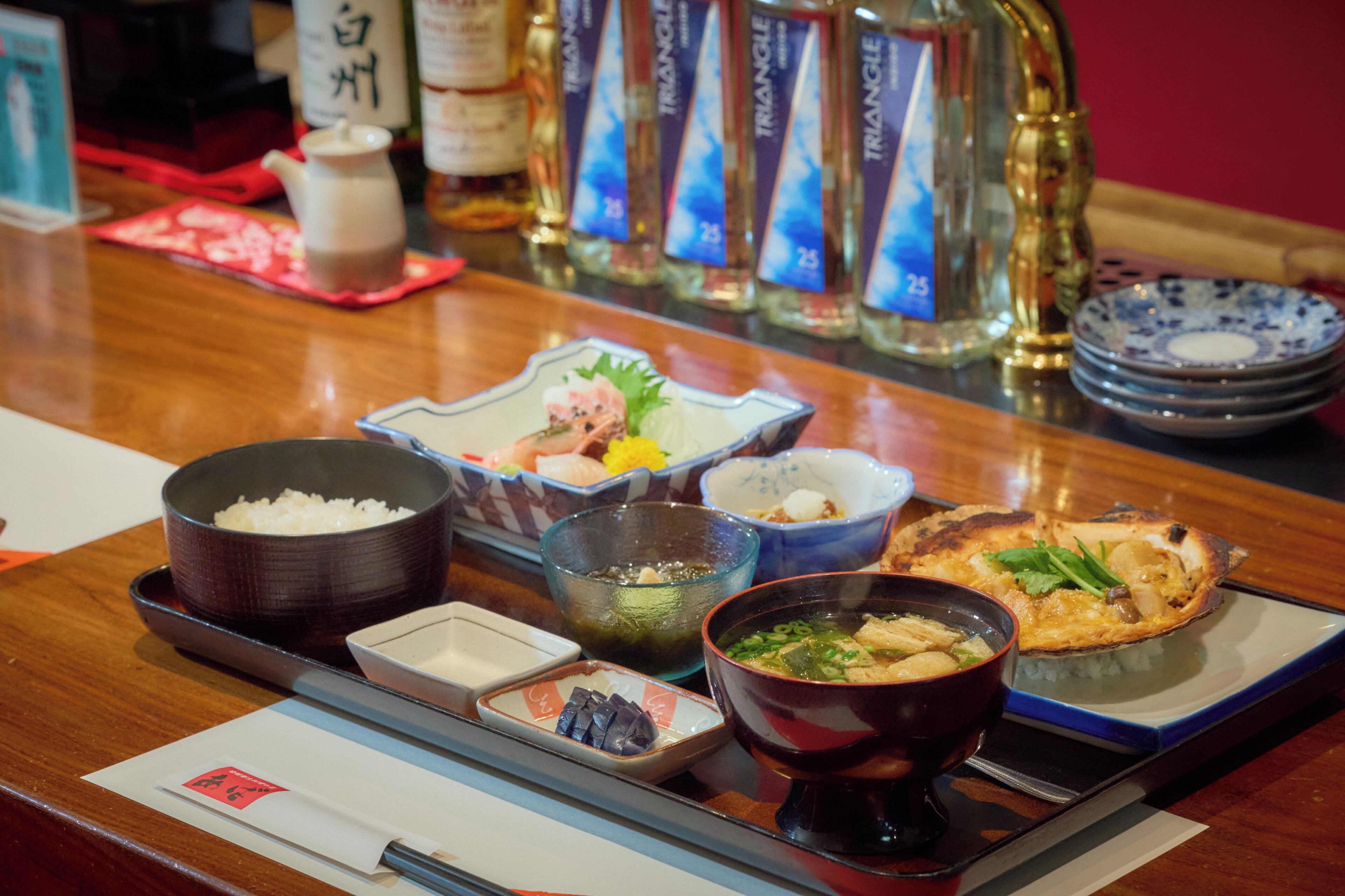 A restaurant counter with various dishes, including rice, soup, and a grilled item, along with condiments and a bottle of liquor.