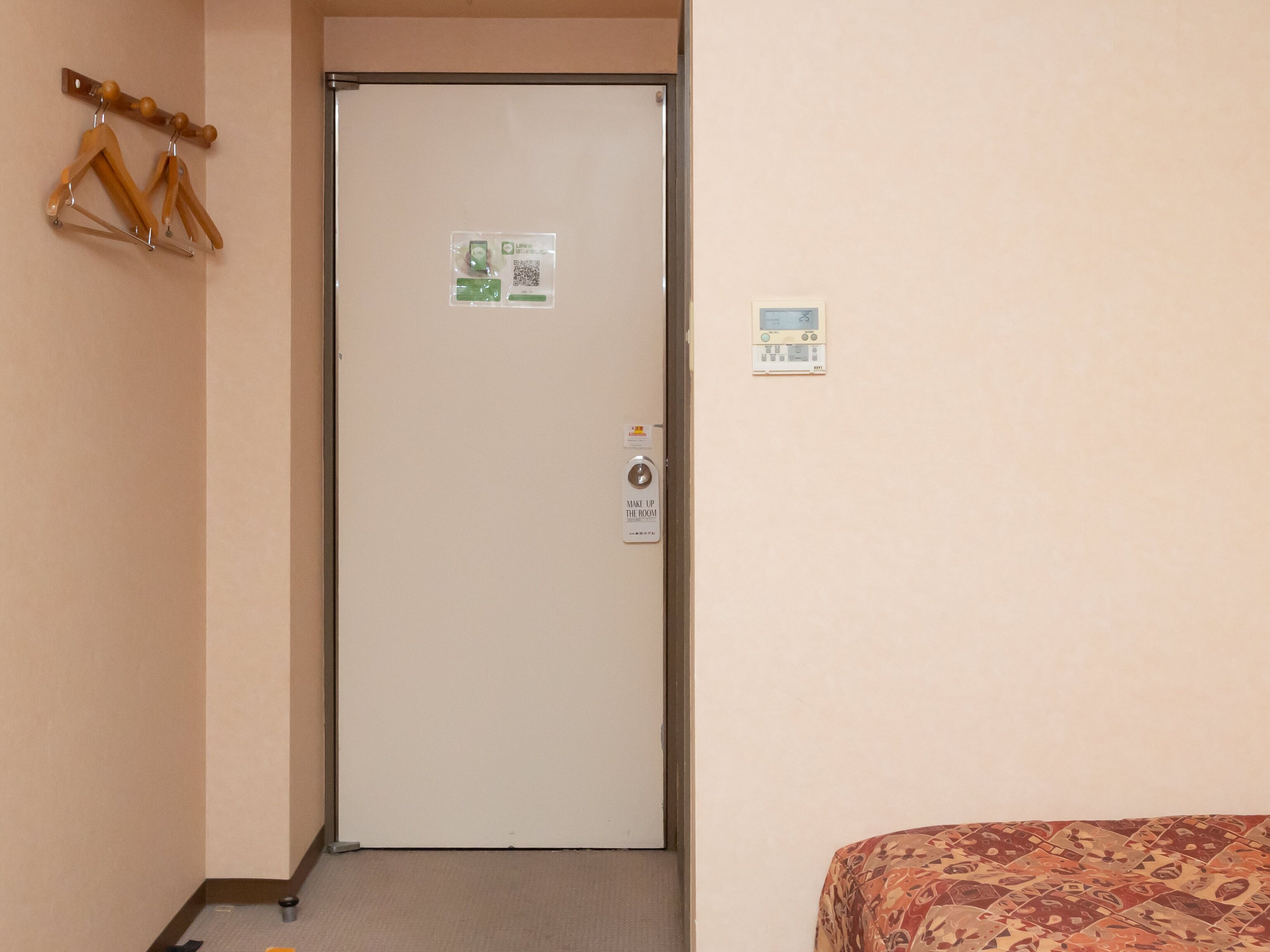 A hotel room with a closed door, a bed with a floral-patterned bedspread, a wall-mounted thermostat, and a coat rack with hangers.
