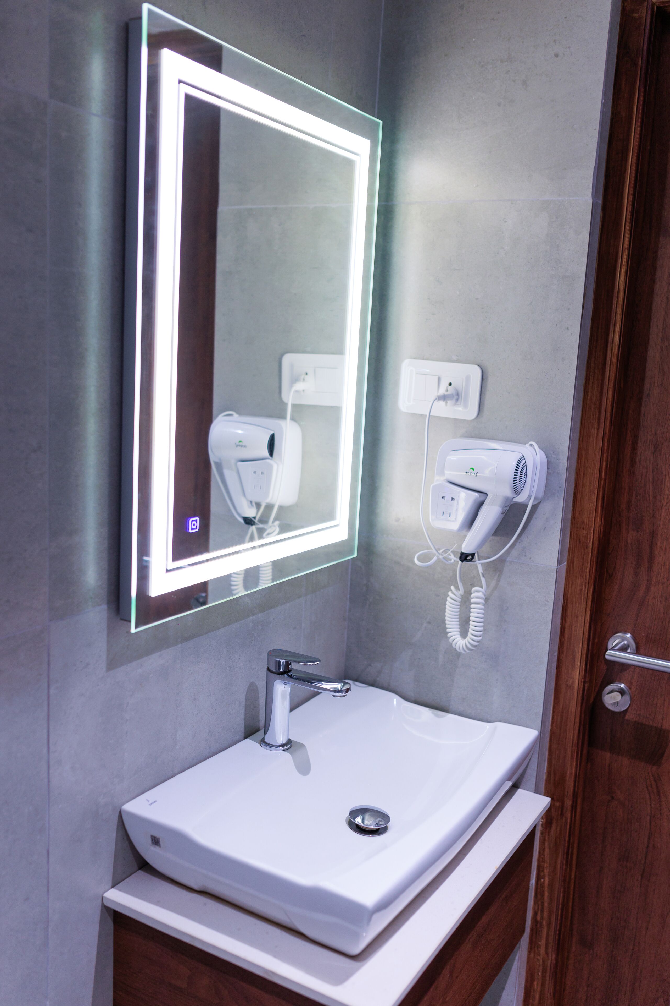 Un baño con tocador moderno, un espejo con iluminación integrada y un secador de pelo montado en la pared.