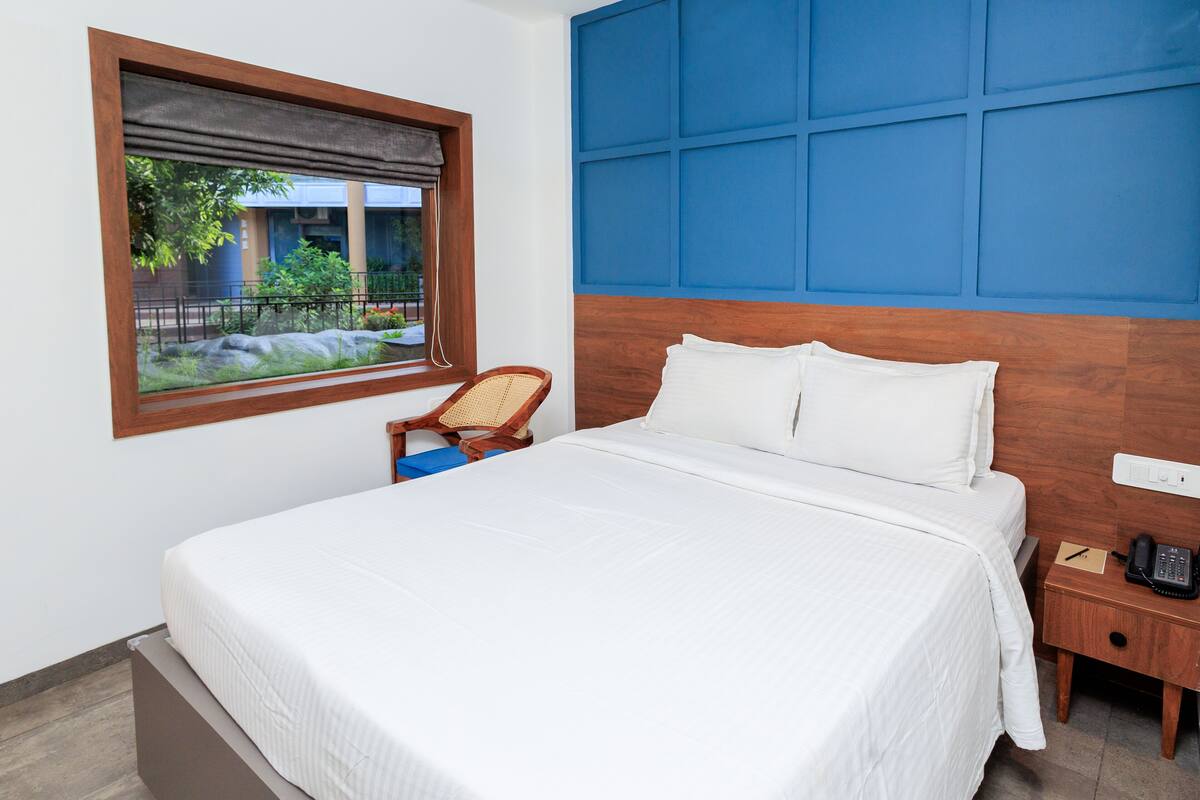 A neatly made bed with white linens, a wooden headboard, a nightstand with a phone, and a window with a view of greenery.