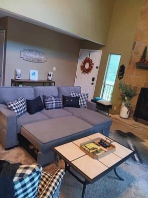 Un salon avec un canapé gris, des coussins à carreaux, une table basse avec des livres et un plateau, une cheminée et une porte décorée d’une guirlande.