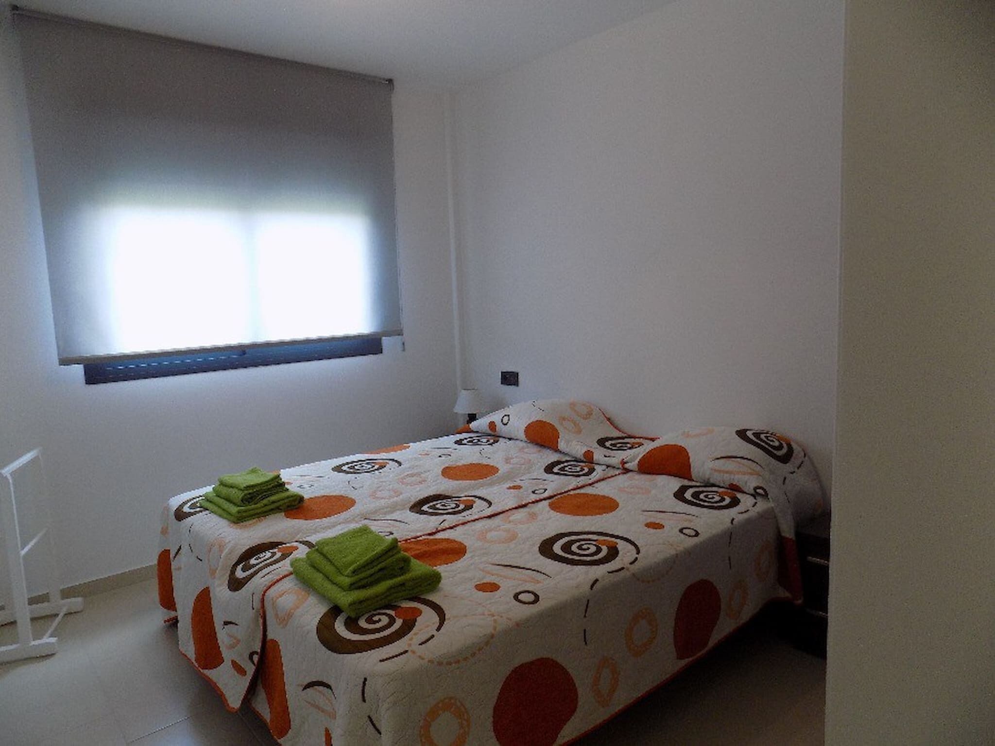 A small, single-bed room with a patterned bedspread, a window with blinds, and a radiator on the wall.