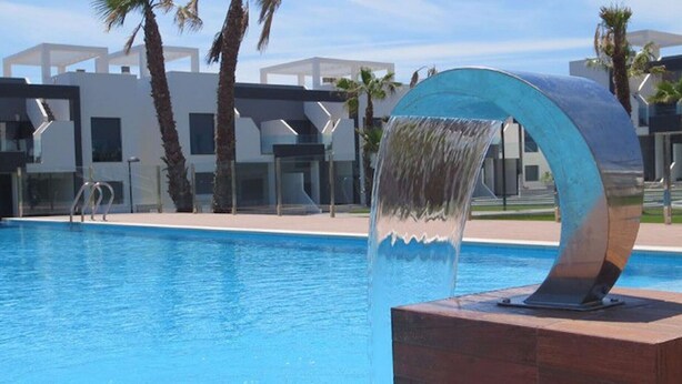 En swimmingpool med ett vattenfall, omgiven av moderna byggnader och palmer.