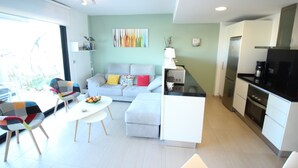 Ein modernes Wohnzimmer mit Sofa, Sesseln und einem Esstisch. Es gibt eine Küchenzeile mit Geräten und einem Kühlschrank.