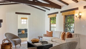 A cozy living room with a sofa, armchairs, and a coffee table. There is a window with a view of greenery outside.