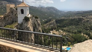 A white building with a red-tiled roof on a rocky outcrop, overlooking a valley with greenery and distant mountains. - Groundfloor Oasis Beach IX Punta Prima (Torrevieja)