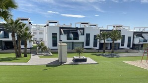 A modern building complex with multiple white structures, a well-maintained lawn, and palm trees. - Groundfloor Oasis Beach IX Punta Prima (Torrevieja)
