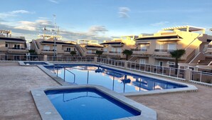 En swimmingpool omgivet af høje bygninger med balkoner, en klar himmel og en byggekran i baggrunden.
