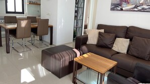 A modern living room with a brown leather sofa, a glass coffee table, and a dining area with a wooden table and chairs.