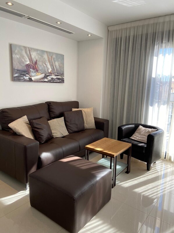 Una sala moderna con un sofá de cuero marrón, una mesa de centro de madera y un sillón negro. Se observa un cuadro de veleros en la pared y una ventana grande con cortinas.