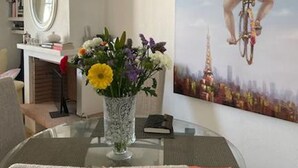 A dining area with a glass table, chairs, a vase of flowers, a painting of a person on a bicycle, and a bookshelf.
