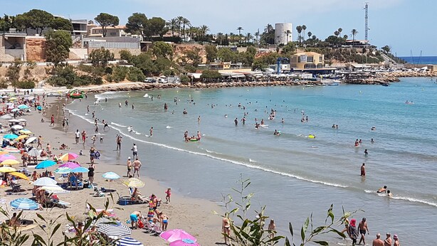 A beach with people swimming and sunbathing, umbrellas, and buildings in the background.