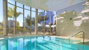 An indoor swimming pool with clear glass walls, a metal ladder, and a view of buildings and trees outside. - Groundfloor GreenHills Villamartin, Orihuela Costa (Villamartín)