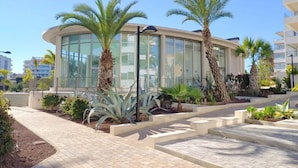 A modern building with large glass windows, surrounded by palm trees and a paved walkway. - Groundfloor GreenHills Villamartin, Orihuela Costa (Villamartín)