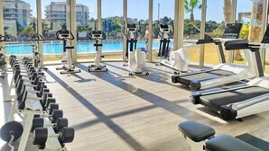 A gym with treadmills, dumbbells, and exercise equipment, overlooking a pool and palm trees. - Groundfloor GreenHills Villamartin, Orihuela Costa (Villamartín)