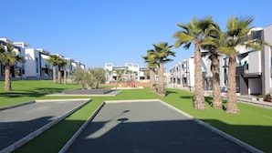 Un hotel moderno con césped verde, palmeras y un parque infantil al fondo.